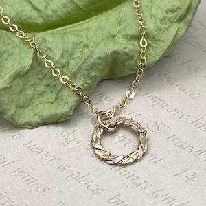 May include: A delicate gold necklace with a circular pendant. The pendant features a braided design with gold and silver tones. The chain is a fine link style, and the necklace is displayed on a leaf.