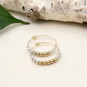 May include: A pair of hoop earrings featuring a thin gold wire frame. Each hoop is accented with a sequence of small, alternating metallic beads in polished gold and textured silver tones, arranged along the lower curve of the circular wire structure.