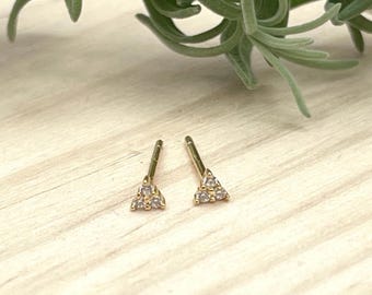 May include: A pair of gold stud earrings. Each earring has a triangle design with small, clear gemstones. The earrings are set against a light-coloured wooden surface, with green foliage in the background.