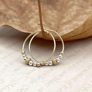 May include: Gold hoop earrings with silver and gold beads. The earrings are presented on a dried leaf, with a blurred background of handwritten text.