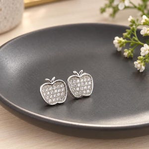 May include: A pair of silver-toned stud earrings shaped like apples, each featuring a textured surface embedded with numerous small, clear, sparkling stones that catch the light, creating a bright and reflective appearance.