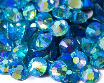 Può includere: Primo piano di un mucchio di strass blu iridescenti. Gli strass sono rotondi e hanno una superficie sfaccettata. Sono sparsi su uno sfondo bianco.