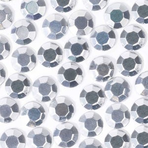 May include: A close-up image of a large quantity of silver-colored, faceted, round rhinestones. The rhinestones are scattered on a white background.