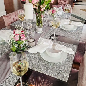 Peut inclure: Une table dressée avec une nappe scintillante argentée, des serviettes blanches et des bougeoirs en verre transparent avec des bougies blanches. Il y a aussi des verres à vin en verre transparent, un vase avec des fleurs roses et un bol de fleurs roses.