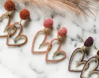 May include: Three pairs of heart-shaped dangle earrings with a beaded design. The earrings are in pink, peach, and purple.
