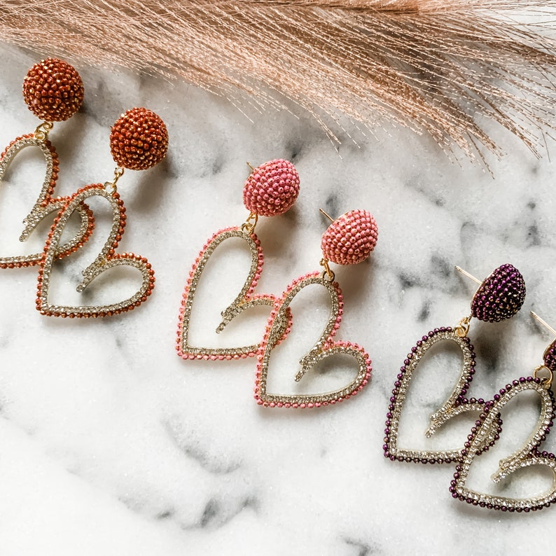 May include: Three pairs of heart-shaped dangle earrings with a beaded design. The earrings are in pink, peach, and purple.