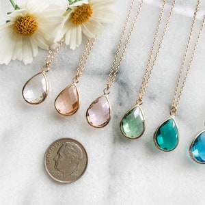 May include: A set of six gold necklaces with teardrop-shaped crystal pendants in various colors, including clear, pink, peach, green, teal, and blue. The necklaces are displayed on a white marble surface.