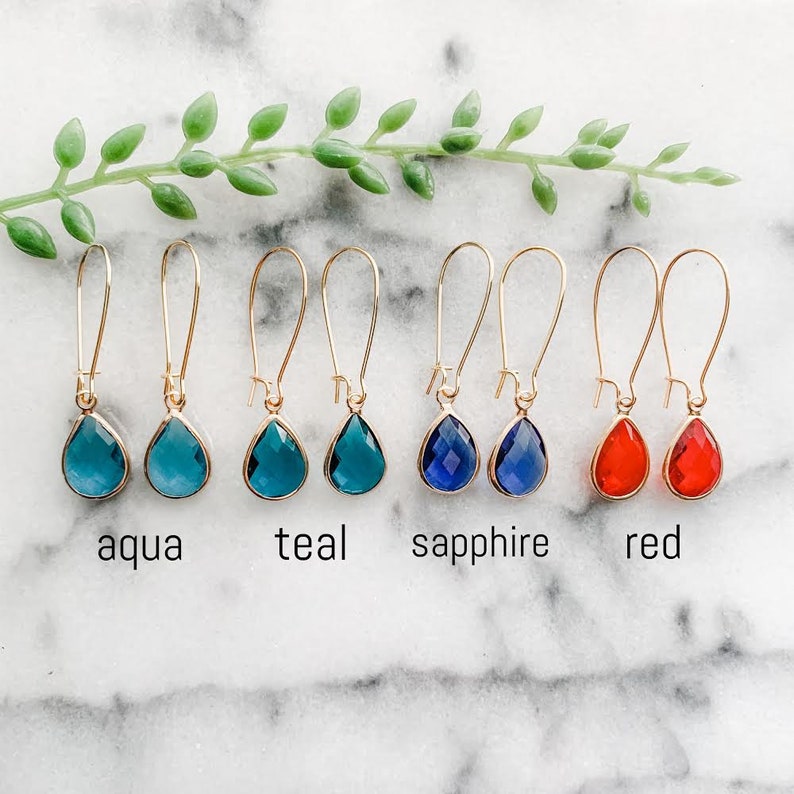 May include: A set of six pairs of gold teardrop earrings with different colored glass stones. The colors are aqua, teal, sapphire, and red.