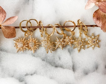 May include: A set of seven gold star-shaped earrings with clear crystal accents. The earrings are hanging from gold hoops.
