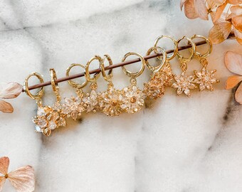 May include: A set of six gold hoop earrings with dangling crystal flower charms. The earrings are displayed on a white marble surface.