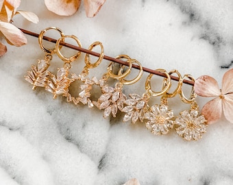 May include: A set of seven gold hoop earrings with dangling crystal flower and leaf charms. The earrings are arranged on a white marble surface.