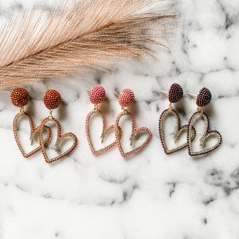 May include: Three pairs of heart-shaped earrings with beaded details. The earrings are in shades of pink, red, and purple.
