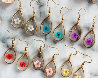 May include: A set of 10 pairs of gold teardrop earrings with pressed flowers inside. The flowers are in various colors, including pink, blue, yellow, and white.