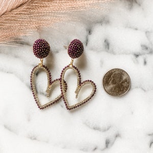 May include: A pair of gold heart-shaped earrings with a burgundy beaded detail. The earrings are dangling and have a burgundy beaded ball at the top.