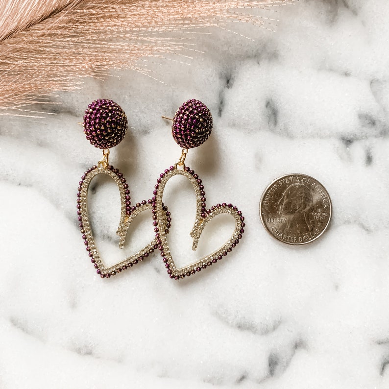 May include: A pair of gold heart-shaped earrings with a burgundy beaded detail. The earrings are dangling and have a burgundy beaded ball at the top.