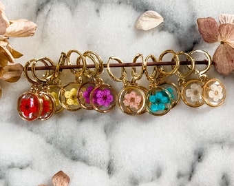 May include: A collection of gold-colored hoop earrings, each featuring a small, round pendant with a colorful dried flower encased in clear resin. The flowers are in various colors, including red, yellow, purple, pink, and blue.