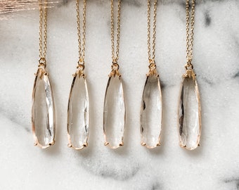 May include: Five gold necklaces with clear teardrop-shaped crystal pendants. The necklaces are arranged in a row on a white marble surface.