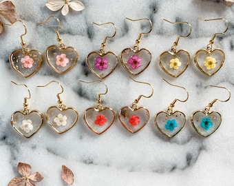 May include: A set of 10 gold heart-shaped earrings with pressed flowers inside. The earrings are arranged in two rows of five. The flowers are in various colors, including pink, yellow, blue, and red.