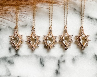 May include: Five gold necklaces with pear-shaped clear crystal pendants surrounded by smaller clear crystals. The necklaces are displayed on a white marble surface.