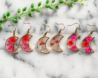 May include: Gold-toned crescent moon earrings filled with dried flowers in shades of red and pink. The earrings have a clear resin center and a delicate gold frame. The earrings are displayed on a white marble surface.