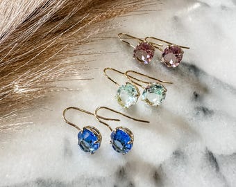 May include: Three pairs of gemstone drop earrings in shades of pink, blue, and green. Each earring features a gold-colored hook and a faceted, round gemstone. The earrings are displayed on a white marble surface.