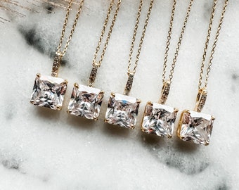 May include: Five gold necklaces with square-shaped clear crystal pendants. The necklaces are arranged in a row on a white marble surface.