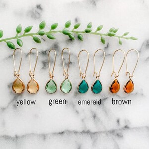May include: Six pairs of gold teardrop earrings with different colored glass stones. The colors are yellow, green, emerald, and brown.