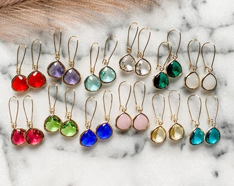May include: A collection of gold-toned drop earrings with teardrop-shaped, faceted gemstones in various colors, including red, purple, green, blue, pink, and clear. The earrings are displayed on a white marble surface.