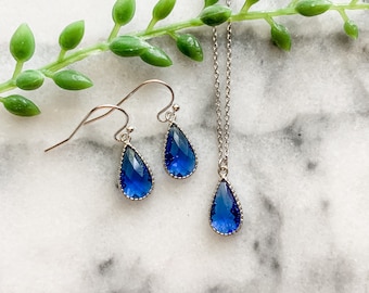 May include: A set of blue teardrop-shaped earrings and a necklace with matching pendants. The jewelry is made of silver and features a delicate, scalloped edge around the blue stones.