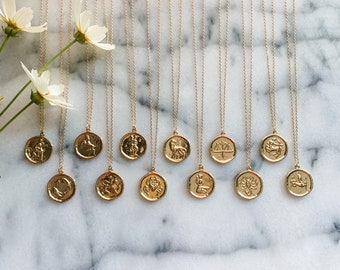 May include: Twelve gold necklaces with zodiac symbols on round pendants. The pendants are arranged in a row on a white marble surface.