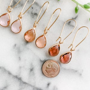 May include: Six pairs of gold teardrop earrings with faceted glass stones in shades of pink and orange. The earrings are displayed on a white marble surface with a US dime for scale.