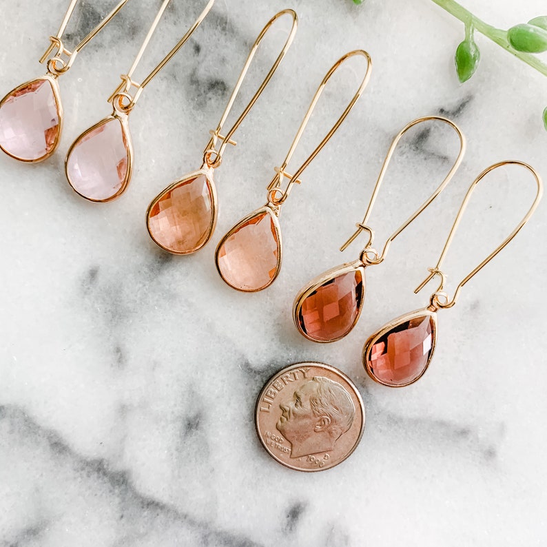 May include: Six pairs of gold teardrop earrings with faceted glass stones in shades of pink and orange. The earrings are displayed on a white marble surface with a US dime for scale.