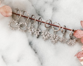 May include: A set of eight silver hoop earrings with crystal accents. The earrings feature a variety of designs, including a circle, a fan, and a leaf.