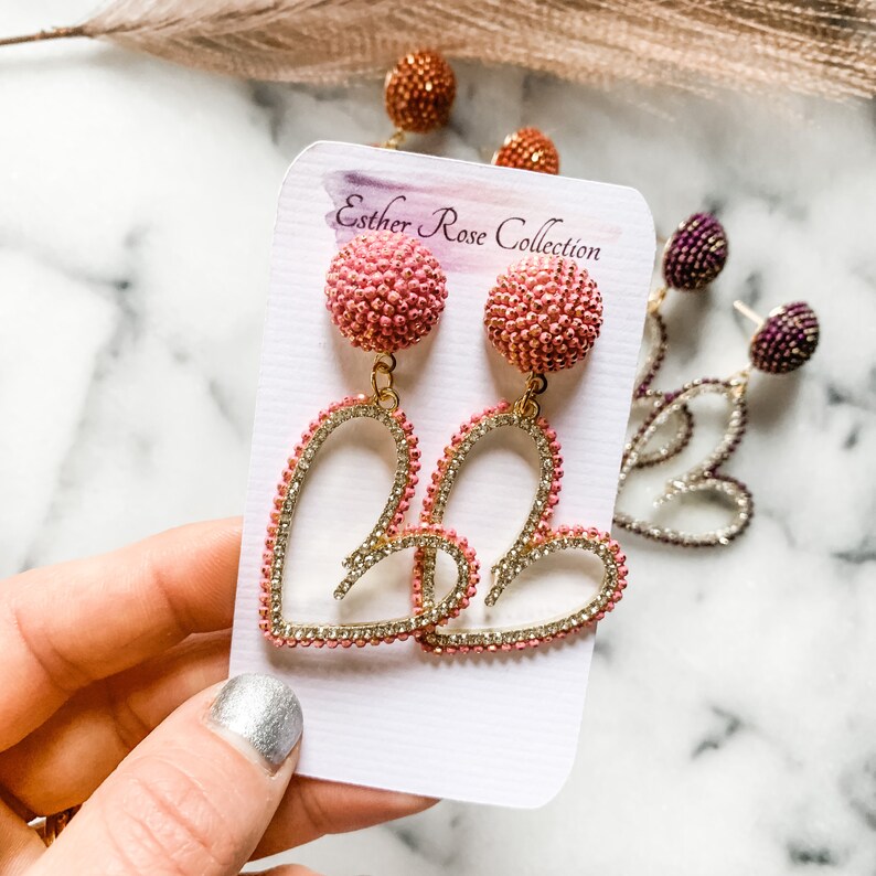 May include: A pair of pink and gold heart-shaped earrings with a beaded detail. The earrings are on a white card with the text "Esther Rose Collection".