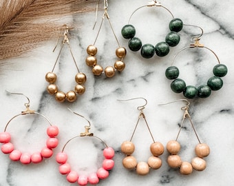 May include: Six pairs of wooden bead earrings in various colors. The earrings are made with round wooden beads and gold-toned metal hoops or teardrop shapes. The colors include green, pink, and natural wood.