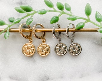 May include: Four gold and silver hoop earrings with a clover charm hanging from each hoop. The earrings are displayed on a gold bar against a white marble background.
