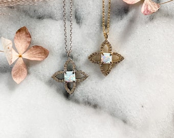 May include: Two necklaces with pendant. One necklace is silver, the other is gold. Each pendant is a four-leaf clover shape with a square opal center stone and small surrounding stones. The necklaces are displayed on a marble surface with dried flowers.