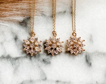 May include: Three gold necklaces with snowflake pendants featuring clear crystals. The necklaces are on a white marble surface.