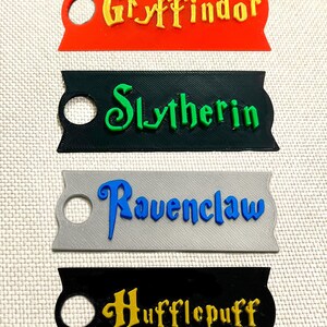 May include: Four rectangular tags in red, black, gray, and black, each with a different house name from the Harry Potter series: Gryffindor, Slytherin, Ravenclaw, and Hufflepuff. Each tag has a hole for attachment.