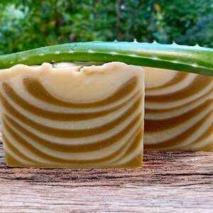 Puede incluir: Dos barras de jab&oacute;n hechas a mano con un patr&oacute;n de remolino blanco y marr&oacute;n, sentadas sobre una superficie de madera con una hoja de aloe vera verde en la parte superior.