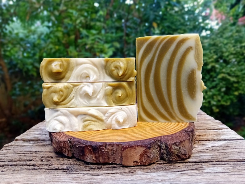 Puede incluir: Cuatro barras de jab&oacute;n artesanal con un dise&ntilde;o de remolino en tonos verdes y blancos. El jab&oacute;n se muestra en una rodaja de madera.