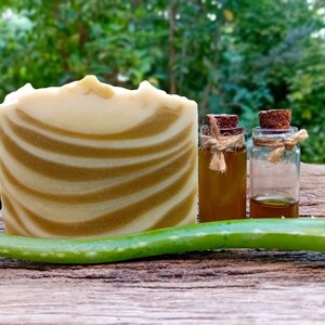 Puede incluir: Una barra de jab&oacute;n blanco y verde en espiral, dos peque&ntilde;os frascos de vidrio con tapas de corcho y una hoja de aloe vera verde sobre una superficie de madera.