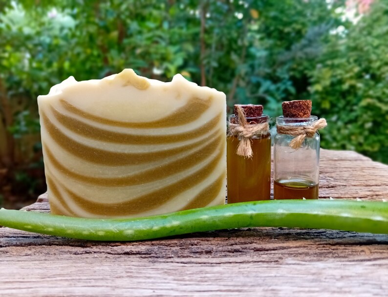 Puede incluir: Una barra de jab&oacute;n blanco y verde en espiral, dos peque&ntilde;os frascos de vidrio con tapas de corcho y una hoja de aloe vera verde sobre una superficie de madera.