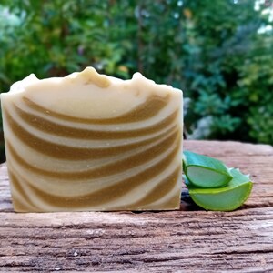 Puede incluir: Una barra de jab&oacute;n artesanal con un patr&oacute;n de remolino blanco y marr&oacute;n. El jab&oacute;n est&aacute; sobre una superficie de madera junto a un trozo de aloe vera verde.