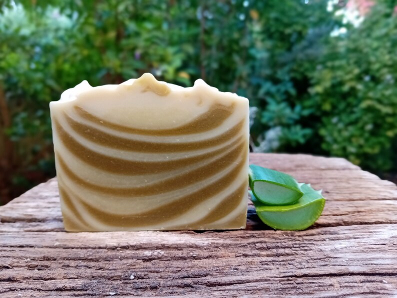 Puede incluir: Una barra de jab&oacute;n artesanal con un patr&oacute;n de remolino blanco y marr&oacute;n. El jab&oacute;n est&aacute; sobre una superficie de madera junto a un trozo de aloe vera verde.