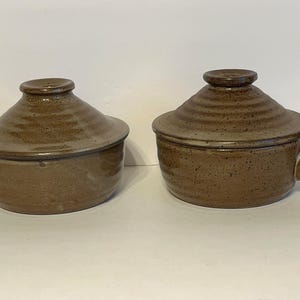 May include: Two brown ceramic casserole dishes with lids and handles. The dishes have a speckled glaze and are perfect for serving individual portions.