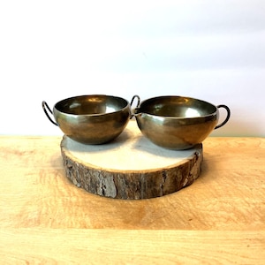 May include: Two vintage brass bowls with black handles, one with a spout, sit on a rustic wooden slice. The bowls have a shiny, metallic finish and are likely used for serving or display. The background is a light wood surface.
