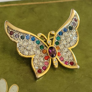 Op de afbeelding: Een gouden vlinder broche met een paarse middelste steen en regenboogkleurige strass steentjes.