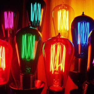 Puede incluir: Un grupo de bombillas de colores con filamentos expuestos, cada una brillando en un color diferente: rojo, naranja, amarillo, verde, azul y morado.