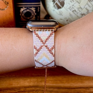 Op de afbeelding: Een Apple Watch in rosé goud met een wit en roze kralenbandje. Het bandje heeft een geometrisch patroon met koperen accenten.